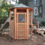 Ein Spielplatz aus Holz mit einer Rutsche, einer Kletterwand, einer Seilbrücke und einer Treppe, der im Freien auf Mulch liegt, mit Bäumen und Gebäuden im Hintergrund.