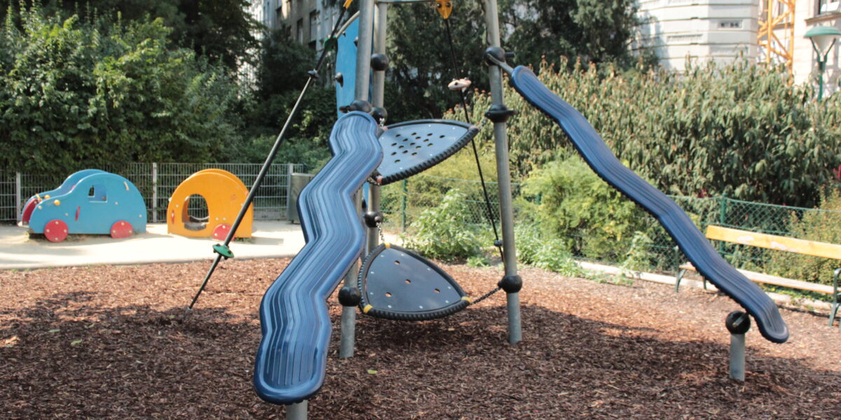 Ein moderner Spielplatz mit zwei blauen Rutschen, Seilelementen und Kletterelementen, umgeben von Holzspänen, Grünpflanzen und einer kleinen tierförmigen Spielstruktur im Hintergrund.