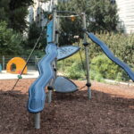 Ein moderner Spielplatz mit zwei blauen Rutschen, Seilelementen und Kletterelementen, umgeben von Holzspänen, Grünpflanzen und einer kleinen tierförmigen Spielstruktur im Hintergrund.
