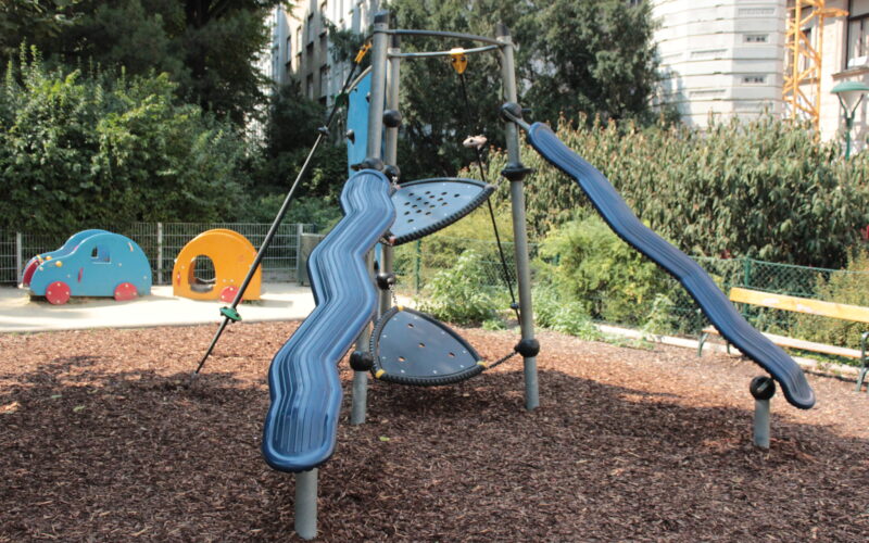 Ein moderner Spielplatz mit zwei blauen Rutschen, Seilelementen und Kletterelementen, umgeben von Holzspänen, Grünpflanzen und einer kleinen tierförmigen Spielstruktur im Hintergrund.