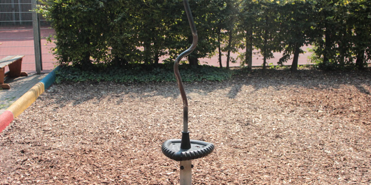 Ein Spielplatz-Drehspielzeug aus Metall mit schwarzem Sockel und rotem Griff steht auf Holzspänen, im Hintergrund sind Büsche und ein Zaun zu sehen.