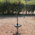 Ein Spielplatz-Drehspielzeug aus Metall mit schwarzem Sockel und rotem Griff steht auf Holzspänen, im Hintergrund sind Büsche und ein Zaun zu sehen.