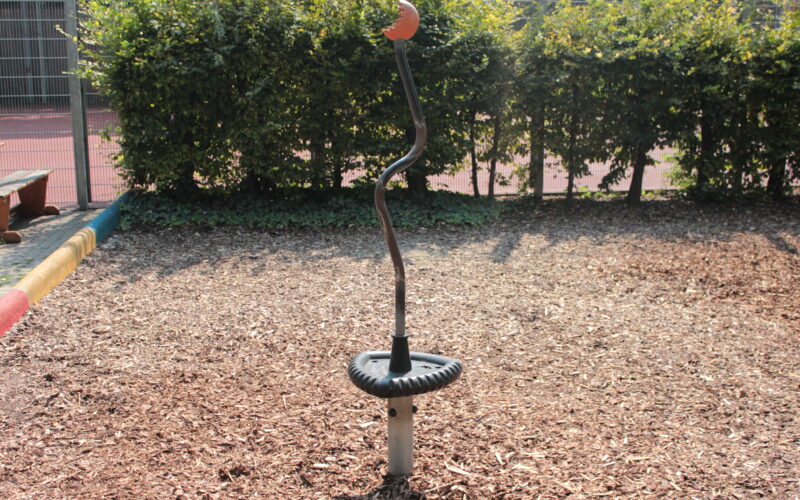 Ein Spielplatz-Drehspielzeug aus Metall mit schwarzem Sockel und rotem Griff steht auf Holzspänen, im Hintergrund sind Büsche und ein Zaun zu sehen.