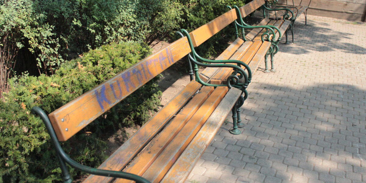 Eine Reihe von leeren Holzbänken mit grünen Metallarmlehnen entlang eines gepflasterten Weges, umgeben von Büschen und Bäumen in einem Park.