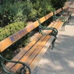 Eine Reihe von leeren Holzbänken mit grünen Metallarmlehnen entlang eines gepflasterten Weges, umgeben von Büschen und Bäumen in einem Park.