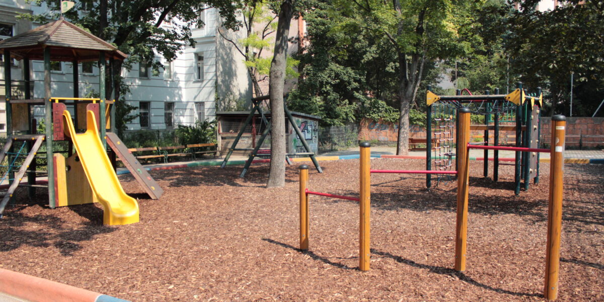 Ein Spielplatz mit einer gelben Rutsche, Klettergerüsten und einem Holzspanbelag, umgeben von Bäumen und nahe gelegenen Gebäuden.