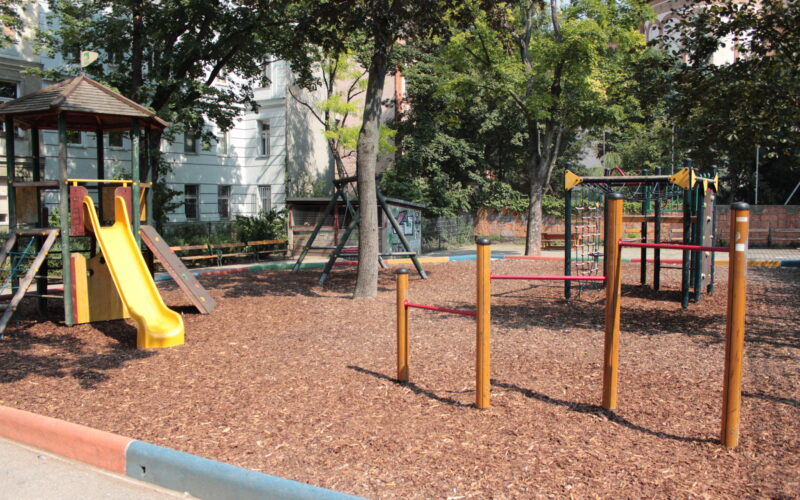 Ein Spielplatz mit einer gelben Rutsche, Klettergerüsten und einem Holzspanbelag, umgeben von Bäumen und nahe gelegenen Gebäuden.