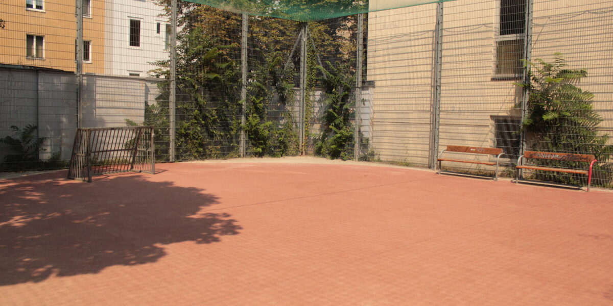 Umzäunter Sportplatz im Freien mit rotem Belag, zwei Bänken, Drahtzaun und Gebäuden in der Nähe im Hintergrund.