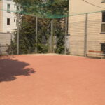 Umzäunter Sportplatz im Freien mit rotem Belag, zwei Bänken, Drahtzaun und Gebäuden in der Nähe im Hintergrund.