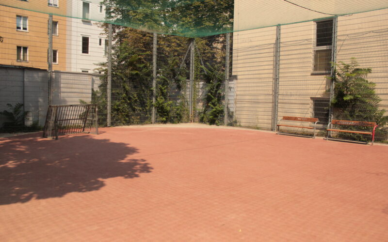 Umzäunter Sportplatz im Freien mit rotem Belag, zwei Bänken, Drahtzaun und Gebäuden in der Nähe im Hintergrund.