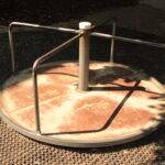 Ein Metallkarussell mit drei Griffen steht auf einer Gummimatte, die teilweise von den Bäumen in der Nähe beschattet wird.