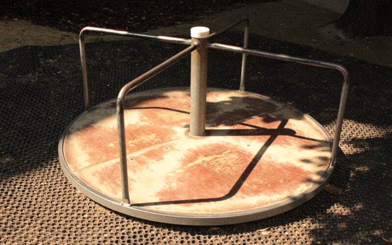 Ein Metallkarussell mit drei Griffen steht auf einer Gummimatte, die teilweise von den Bäumen in der Nähe beschattet wird.