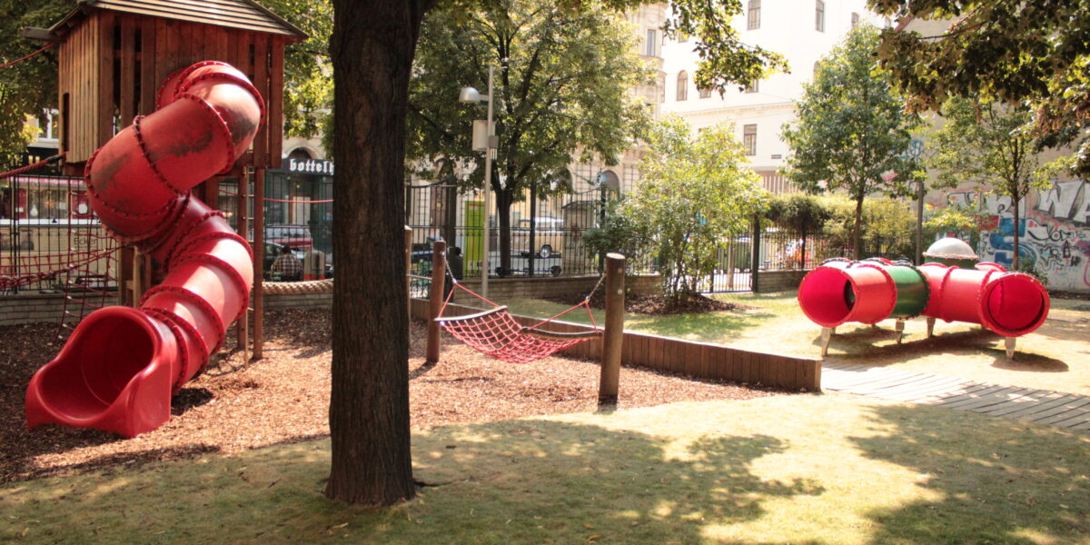Ein Spielplatz mit einer roten Röhrenrutsche, einem Seilkletternetz und einer roten Tunnelkonstruktion, umgeben von Bäumen und einer Rasenfläche.