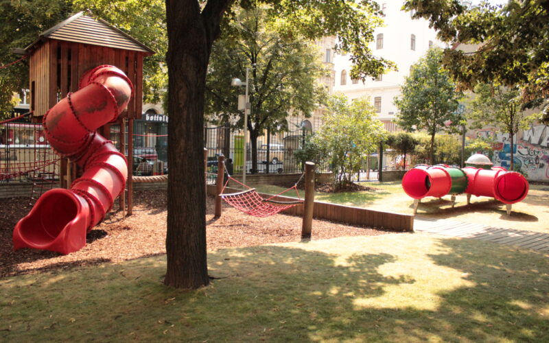 Ein Spielplatz mit einer roten Röhrenrutsche, einem Seilkletternetz und einer roten Tunnelkonstruktion, umgeben von Bäumen und einer Rasenfläche.