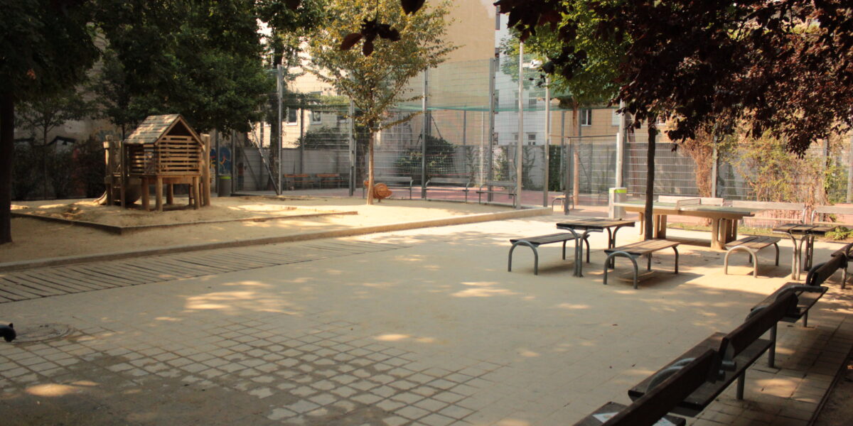 Ein ruhiger Spielplatz im Freien mit Bänken, Picknicktischen, einem Holzspielhaus und einem eingezäunten Sportplatz im Hintergrund.