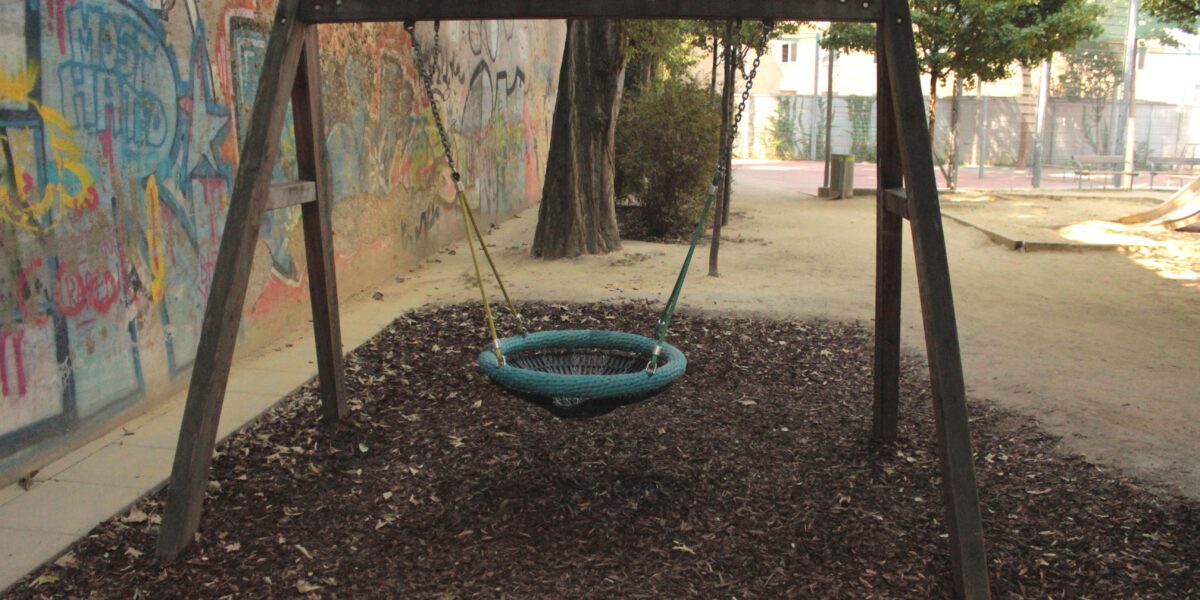 Ein Spielplatz mit einer runden Netzschaukel, die an einem Holzrahmen aufgehängt ist, umgeben von Mulch und mit Graffiti beschmierten Wänden im Hintergrund.