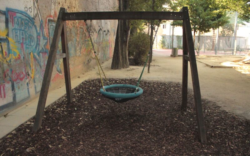 Ein Spielplatz mit einer runden Netzschaukel, die an einem Holzrahmen aufgehängt ist, umgeben von Mulch und mit Graffiti beschmierten Wänden im Hintergrund.