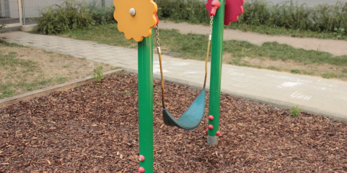 Eine kleine einsitzige Schaukel mit einem flexiblen schwarzen Sitz ist zwischen zwei grünen Pfosten mit roten und gelben zahnradförmigen Aufsätzen auf einem Spielplatz mit Holzspänen aufgehängt.