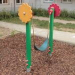 Eine kleine einsitzige Schaukel mit einem flexiblen schwarzen Sitz ist zwischen zwei grünen Pfosten mit roten und gelben zahnradförmigen Aufsätzen auf einem Spielplatz mit Holzspänen aufgehängt.
