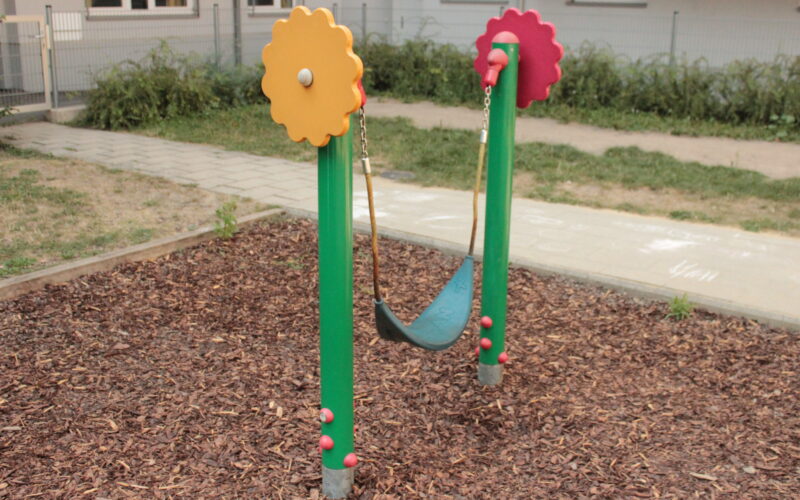 Eine kleine einsitzige Schaukel mit einem flexiblen schwarzen Sitz ist zwischen zwei grünen Pfosten mit roten und gelben zahnradförmigen Aufsätzen auf einem Spielplatz mit Holzspänen aufgehängt.