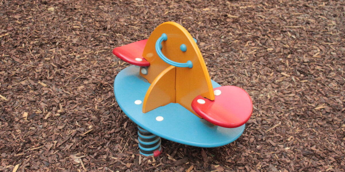 Bunte Spielplatz Feder Reiter mit zwei roten Sitzen, eine gelbe Mittelplatte mit einem Smiley-Gesicht, und eine blaue Basis, auf Holzspan Boden gesetzt.