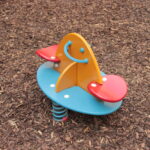 Bunte Spielplatz Feder Reiter mit zwei roten Sitzen, eine gelbe Mittelplatte mit einem Smiley-Gesicht, und eine blaue Basis, auf Holzspan Boden gesetzt.