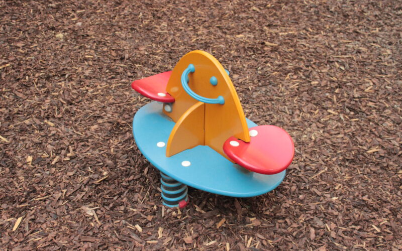 Bunte Spielplatz Feder Reiter mit zwei roten Sitzen, eine gelbe Mittelplatte mit einem Smiley-Gesicht, und eine blaue Basis, auf Holzspan Boden gesetzt.