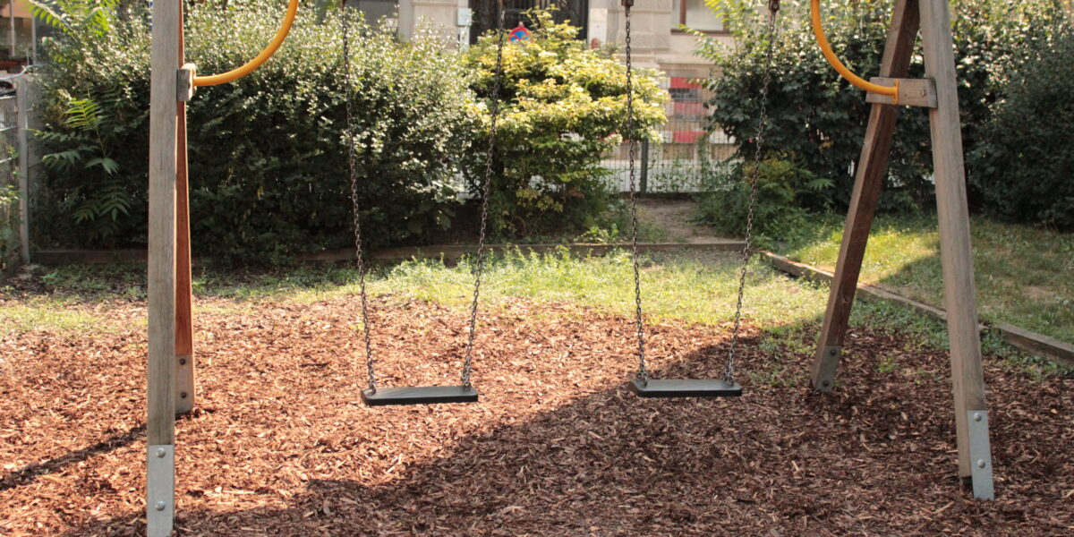 Ein hölzernes Schaukel-Set mit zwei leeren Schaukeln hängt über dem Mulch auf einem Spielplatz im Freien, umgeben von Grün und Büschen.