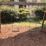 Ein hölzernes Schaukel-Set mit zwei leeren Schaukeln hängt über dem Mulch auf einem Spielplatz im Freien, umgeben von Grün und Büschen.