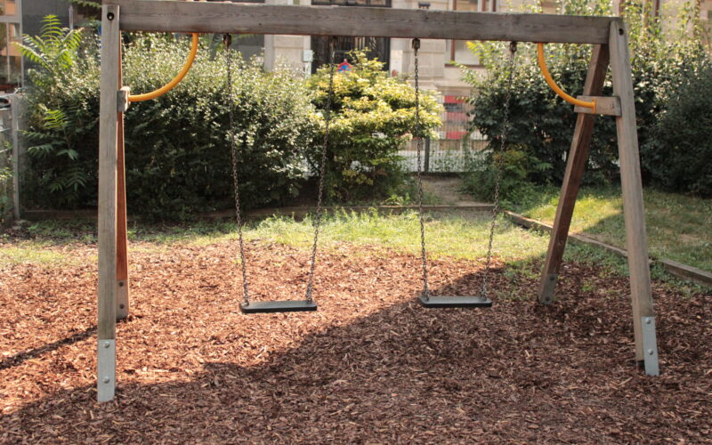 Ein hölzernes Schaukel-Set mit zwei leeren Schaukeln hängt über dem Mulch auf einem Spielplatz im Freien, umgeben von Grün und Büschen.