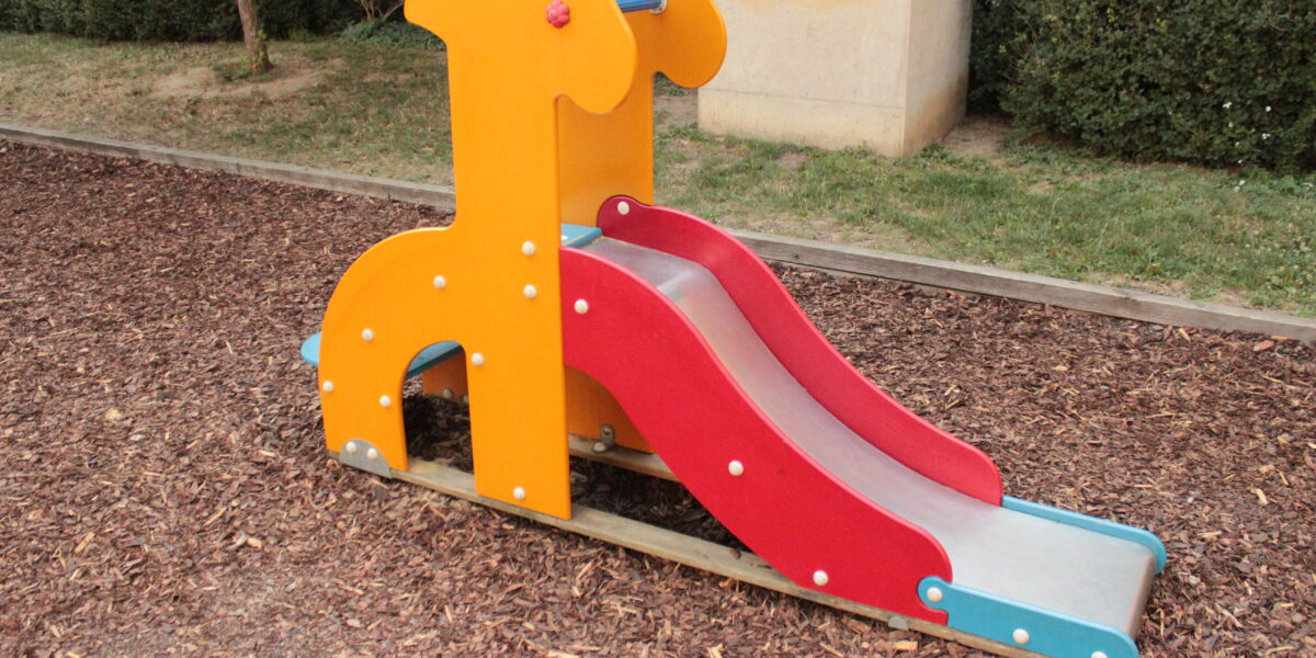Eine kleine Spielplatzrutsche in Form einer Giraffe, mit orangefarbenen Seiten und einer roten Rutschfläche, die auf einem mit Mulch bedeckten Boden steht.