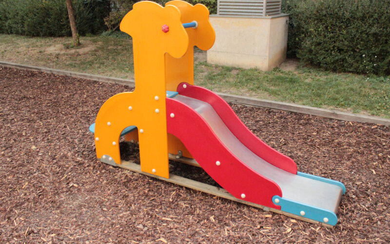 Eine kleine Spielplatzrutsche in Form einer Giraffe, mit orangefarbenen Seiten und einer roten Rutschfläche, die auf einem mit Mulch bedeckten Boden steht.