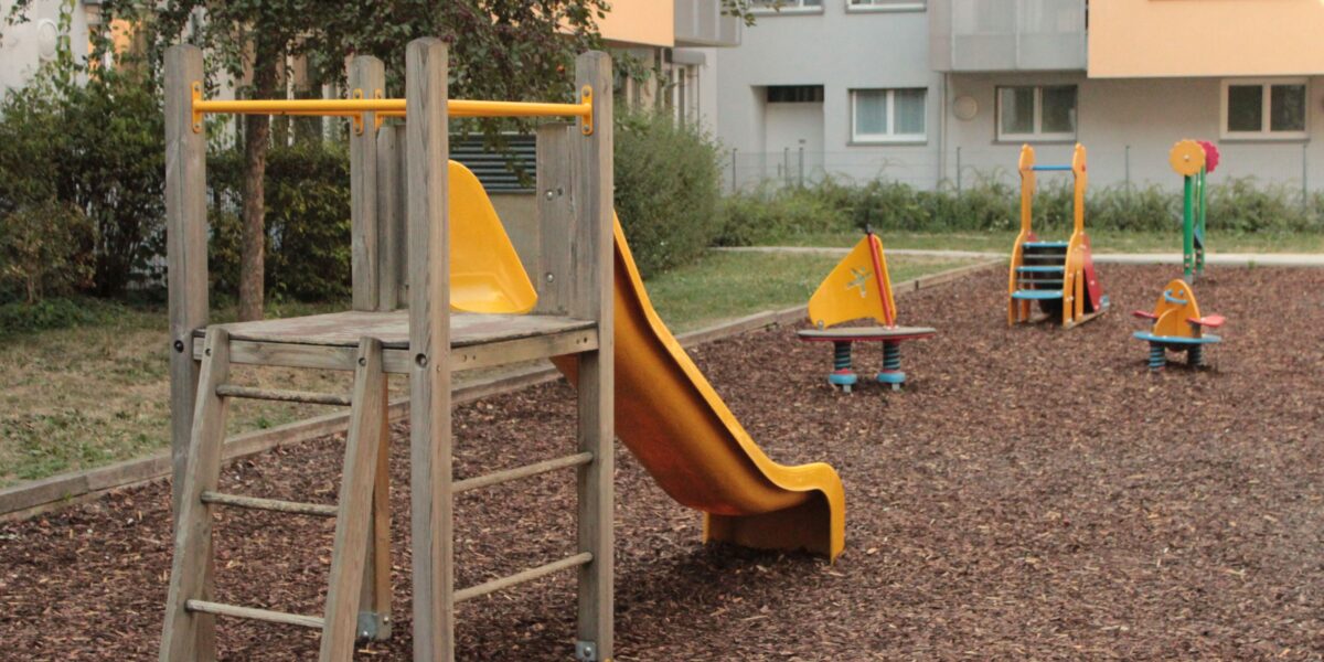 Ein kleiner Spielplatz mit einer Holzrutsche und mehreren Springreitern auf Holzspanboden in der Nähe von Wohnhäusern.