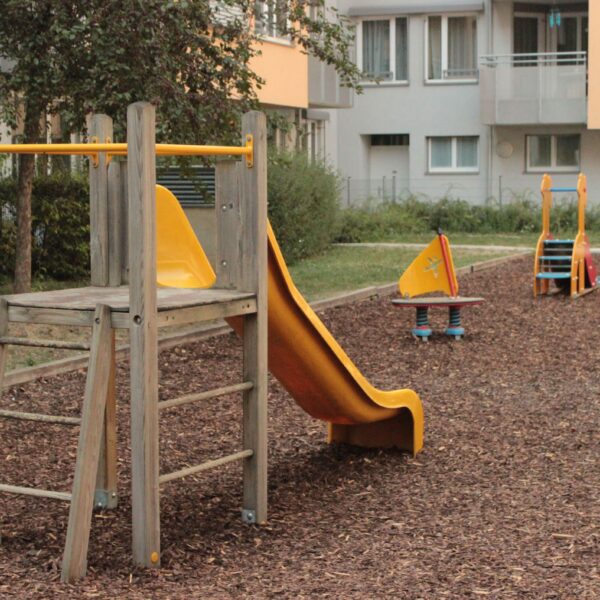 Ein kleiner Spielplatz mit einer Holzrutsche und mehreren Springreitern auf Holzspanboden in der Nähe von Wohnhäusern.
