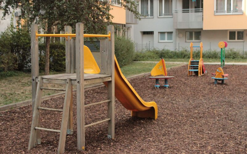 Ein kleiner Spielplatz mit einer Holzrutsche und mehreren Springreitern auf Holzspanboden in der Nähe von Wohnhäusern.