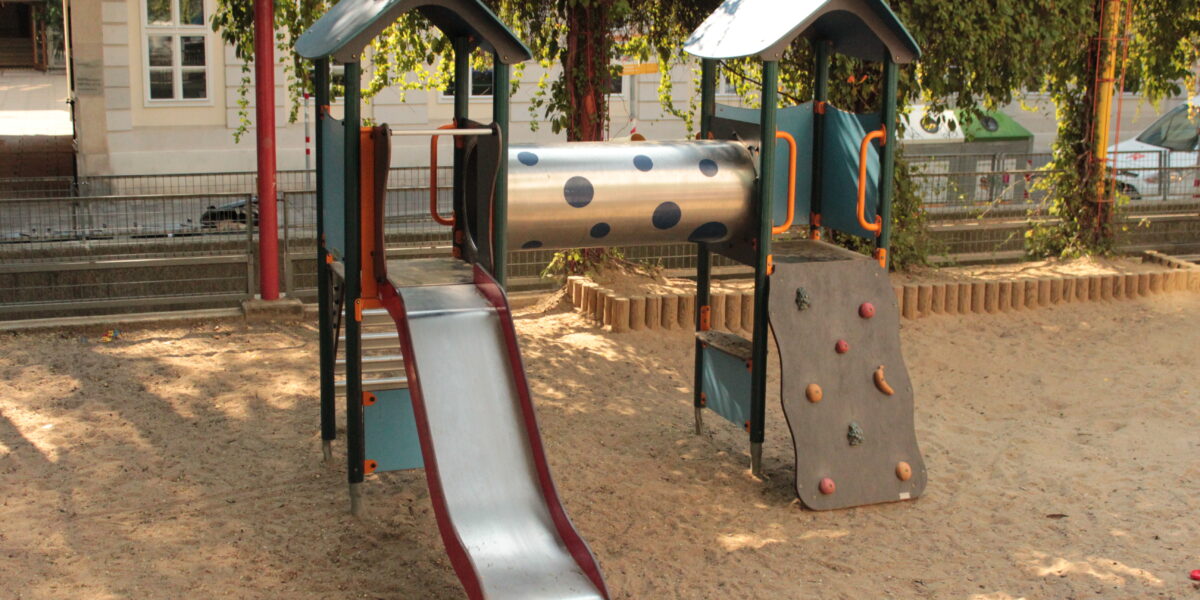 Spielplatz mit einer Rutsche, einer Kletterwand und einem Tunnel auf Sand mit Bäumen und einem Zaun im Hintergrund.