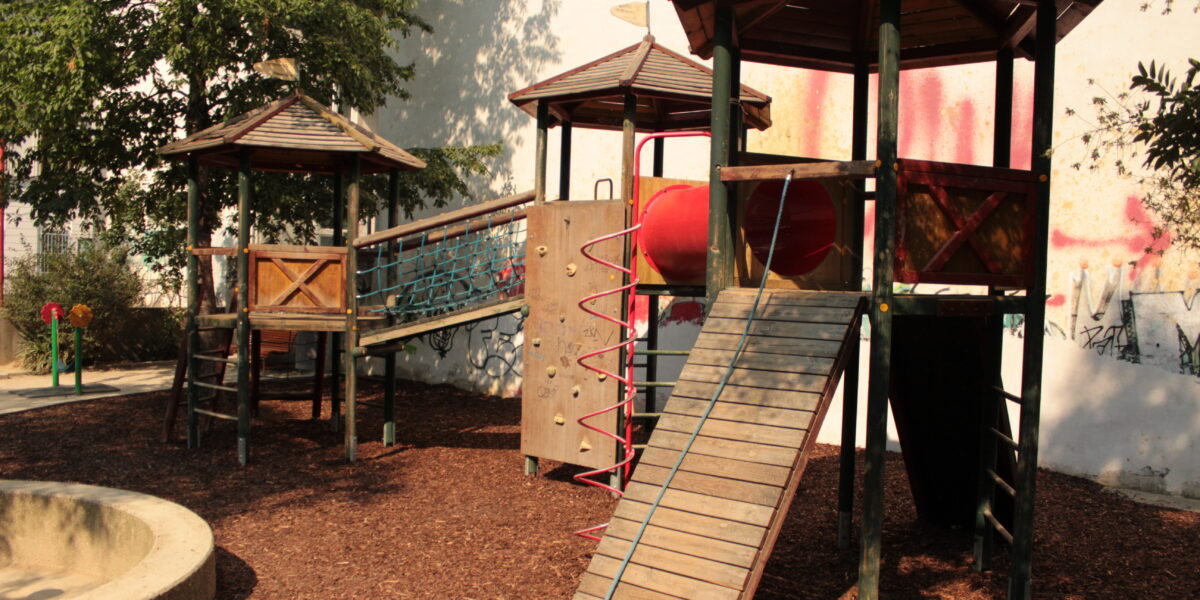 Spielplatz im Freien mit Klettergerüsten, Rutschen und Brücken aus Holz, umgeben von Bäumen und Mulch, neben einer weißen Wand mit Graffiti.