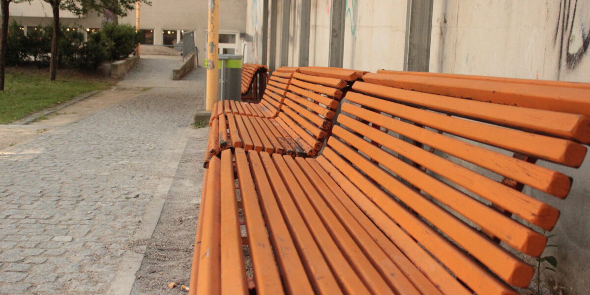 Orangefarbene Holzbänke stehen auf einem Kopfsteinpflasterweg neben einem Gebäude mit Graffiti, in dessen Nähe Bäume und ein Mülleimer stehen.