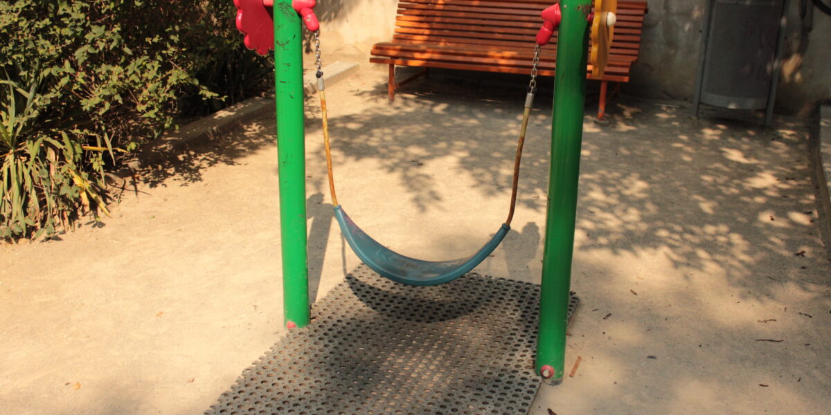Eine kleine leere Schaukel mit einem flexiblen blauen Sitz hängt zwischen zwei grünen Pfosten in einem sonnigen Park, mit einer Holzbank und einem Mülleimer im Hintergrund.
