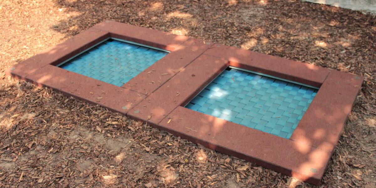 Zwei rechteckige, in den Boden eingelassene Trampoline mit blauen gewebten Oberflächen und roten Rahmen befinden sich in einem mit Mulch bedeckten Spielplatzbereich.