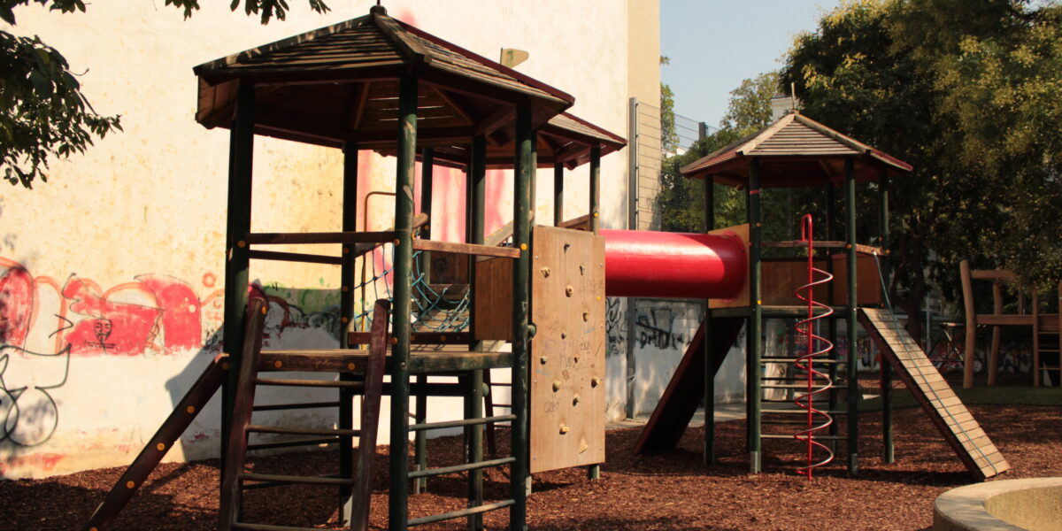 Spielplatz mit zwei hölzernen Spielgeräten, die durch eine rote Tunnelrutsche, Kletterelemente und Mulchbodenbelag verbunden sind, mit Bäumen und Graffiti an einer Wand im Hintergrund.