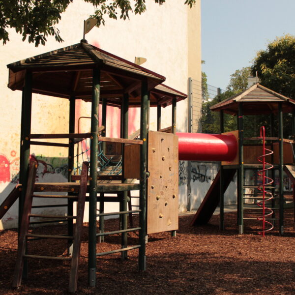 Spielplatz mit zwei hölzernen Spielgeräten, die durch eine rote Tunnelrutsche, Kletterelemente und Mulchbodenbelag verbunden sind, mit Bäumen und Graffiti an einer Wand im Hintergrund.