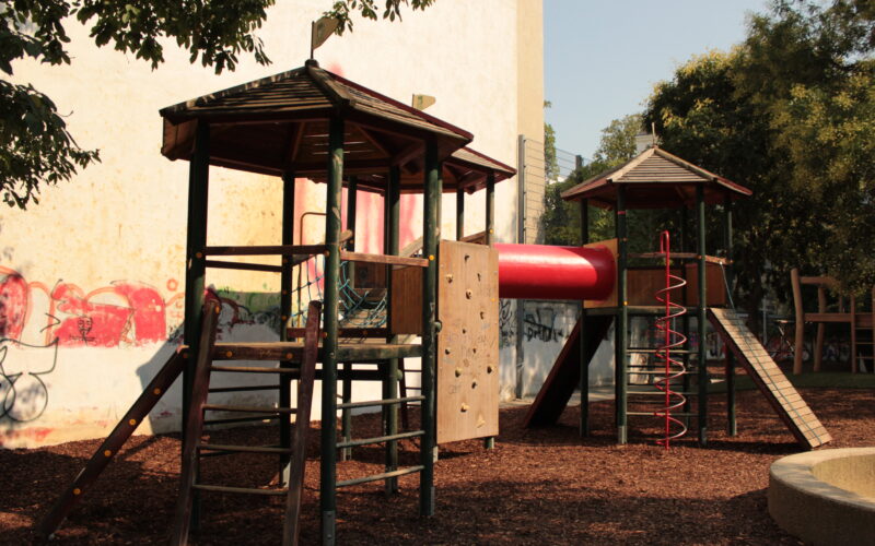 Spielplatz mit zwei hölzernen Spielgeräten, die durch eine rote Tunnelrutsche, Kletterelemente und Mulchbodenbelag verbunden sind, mit Bäumen und Graffiti an einer Wand im Hintergrund.