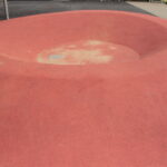 Eine große, flache, schüsselförmige Vertiefung aus rotem Gummi im Boden, wahrscheinlich Teil eines Spielplatzes oder einer städtischen Parkanlage.