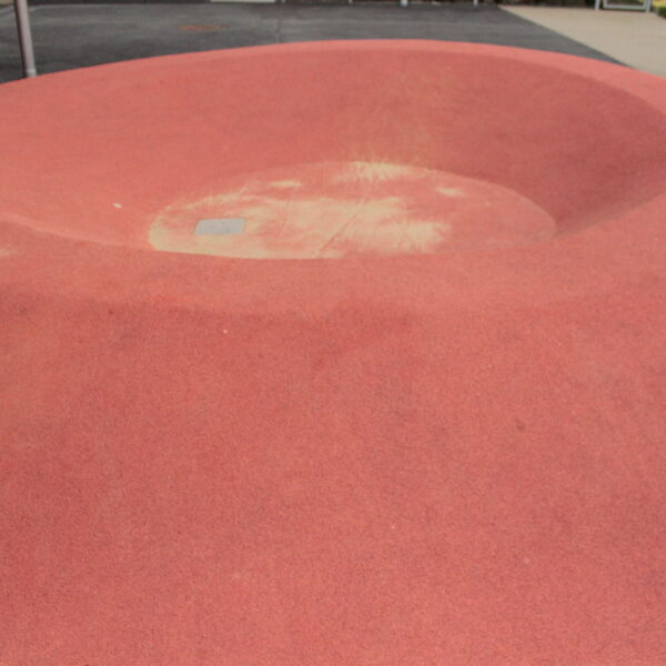Eine große, flache, schüsselförmige Vertiefung aus rotem Gummi im Boden, wahrscheinlich Teil eines Spielplatzes oder einer städtischen Parkanlage.