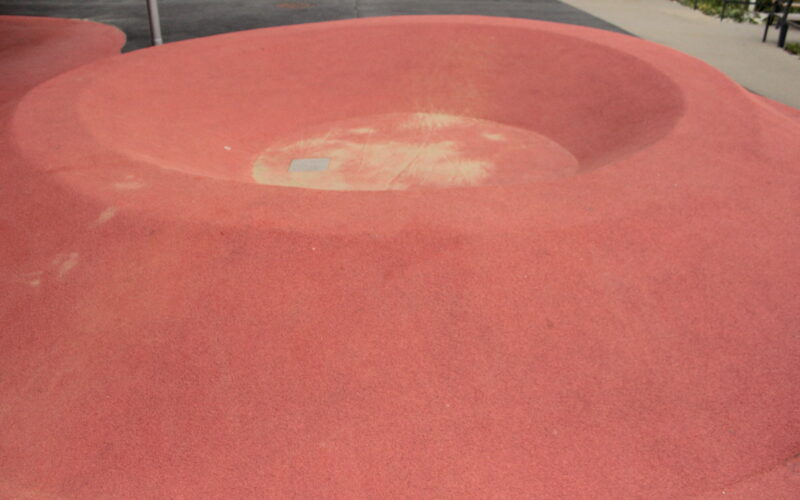 Eine große, flache, schüsselförmige Vertiefung aus rotem Gummi im Boden, wahrscheinlich Teil eines Spielplatzes oder einer städtischen Parkanlage.