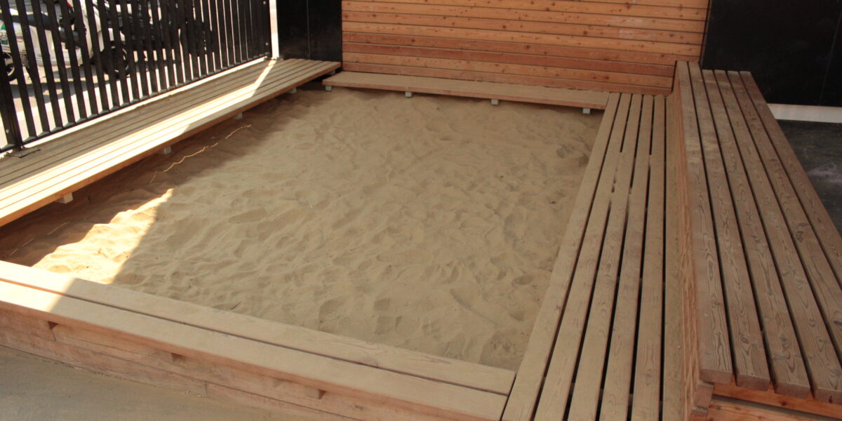 Ein rechteckiger hölzerner Sandkasten mit Bänken an drei Seiten, der mit Sand gefüllt ist und im Freien in der Nähe eines Gebäudes und eines schwarzen Metallzauns steht.