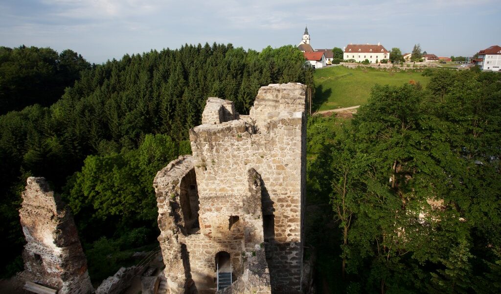 Ruinen eines alten Steingebäudes, umgeben von dichten grünen Bäumen, im Hintergrund sind unter einem blauen Himmel ein Dorf und eine Kirche zu sehen.