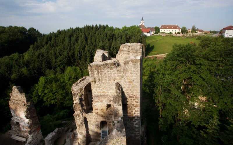 Ruinen eines alten Steingebäudes, umgeben von dichten grünen Bäumen, im Hintergrund sind unter einem blauen Himmel ein Dorf und eine Kirche zu sehen.