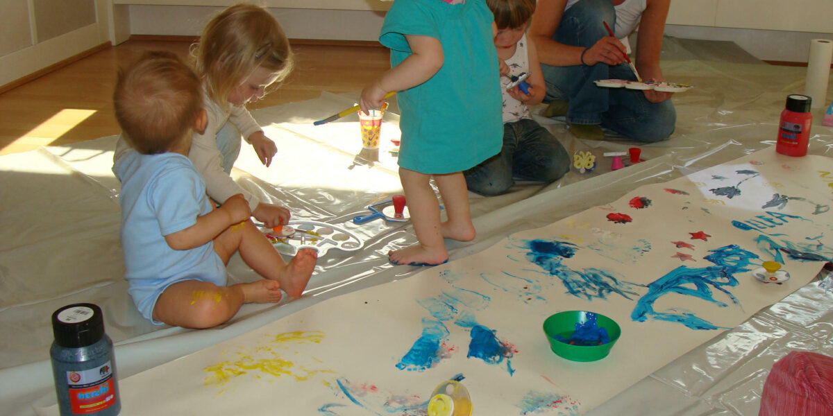 Mehrere kleine Kinder und ein Erwachsener sitzen auf dem Boden um ein großes Blatt Papier herum und malen mit den Fingern in verschiedenen Farben. Künstlerbedarf und Farbflaschen liegen verstreut in der Nähe.